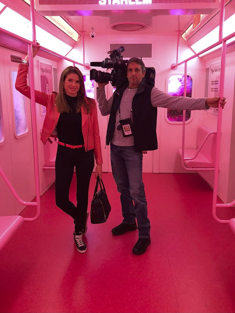 Jen anchoring in the field with her cameraman inside a subway car decked out in neon pink lighting, wearing black jeans, a black top, black sneakers, a pink belt and pink leather jacket.
