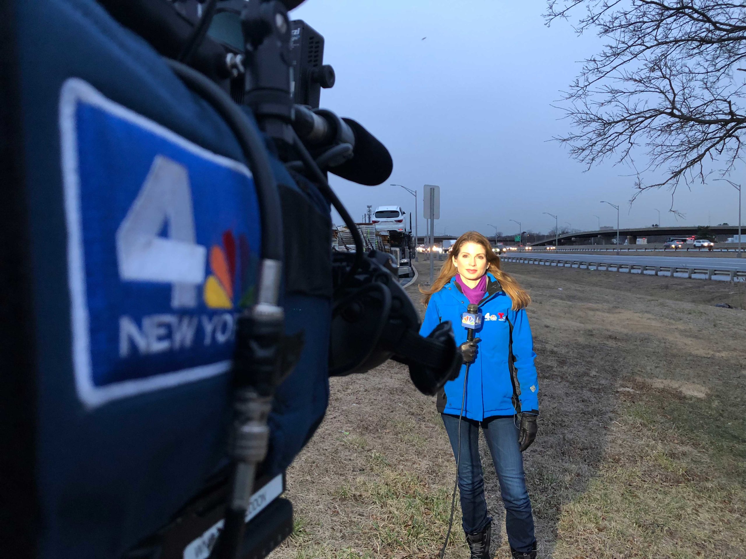 Jen on the field in front of a throughway, wearing an NBC 4 New York winter jacket. Picture is taken from behind a news camera with the NBC 4 New York logo on the side.